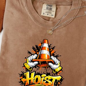May include: Brown t-shirt with a graphic of a traffic cone held by gloved hands, the word "Hoist" in yellow and orange. The shirt has a Comfort Colors tag and a silver necklace.