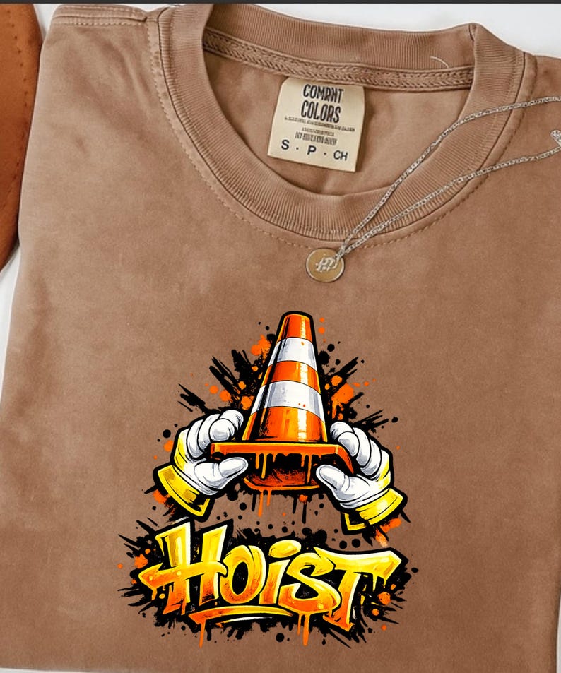 May include: Brown t-shirt with a graphic of a traffic cone held by gloved hands, the word "Hoist" in yellow and orange. The shirt has a Comfort Colors tag and a silver necklace.