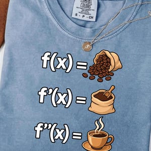 Op de afbeelding: Een lichtblauw t-shirt met ronde hals en een grafische print van drie wiskundige afgeleide vergelijkingen. De eerste regel toont f(x) gelijk aan een zak koffiebonen, de tweede f'(x) gelijk aan gemalen koffie en de derde f''(x) gelijk aan een kopje koffie.