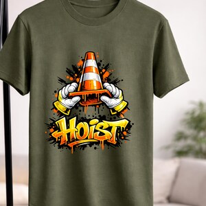 May include: Olive green t-shirt featuring a graphic of a traffic cone held by cartoon hands with yellow gloves. The word "Hoist" is written in orange graffiti-style lettering below the cone.