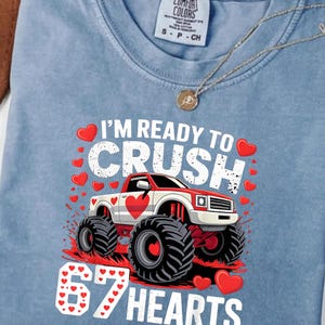 Peut inclure: T-shirt bleu clair avec un motif de camion monstre rouge et blanc avec des cœurs et le texte "I'M READY TO CRUSH 67 HEARTS". Le t-shirt a une étiquette Comfort Colors.