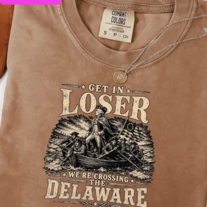 Peut inclure: Un t-shirt Comfort Colors beige avec un motif d'un bateau traversant une rivière, avec le texte "GET IN LOSER WE'RE CROSSING THE DELAWARE". Un collier argenté avec un pendentif est sur le t-shirt. Le t-shirt a le texte "TRENDING DESIGNS" et "PNG & SVG".