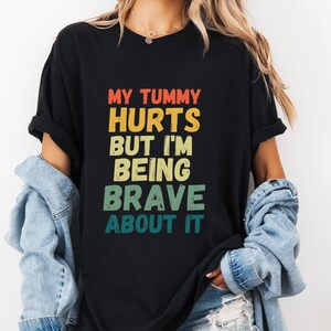Peut inclure: T-shirt noir avec le texte "MY TUMMY HURTS BUT I'M BEING BRAVE ABOUT IT" en rouge, jaune, vert et bleu sarcelle. Le t-shirt est porté avec une veste en jean et un short.