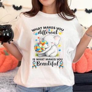 Peut inclure: T-shirt blanc avec le texte "CE QUI VOUS REND DIFFÉRENT EST CE QUI VOUS REND BEAU." Le t-shirt présente un éléphant de dessin animé étreignant une pièce de puzzle. Des étoiles colorées et des rubans de sensibilisation à l'autisme sont également présents.