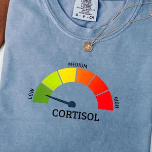 May include: A light blue t-shirt with a graphic of a cortisol level gauge. The gauge ranges from green (low) to red (high), with the word "CORTISOL" below. A gold necklace with a pendant is also visible.