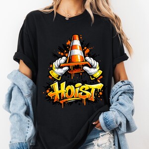 May include: Black t-shirt featuring a graphic of a traffic cone held by gloved hands, with the word "Hoist" in orange and yellow graffiti style. The design includes orange and black paint splatter effects.