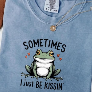 以下が含まれることがあります： 緑色のカエルグラフィックと黒い文字「SOMETIMES I just BE KISSIN'」がプリントされた、ライトブルーのComfort Colors Tシャツ。カエルは大きな目を持ち、小さな赤いハートに囲まれています。金のネックレスがシャツの上にかけられています。