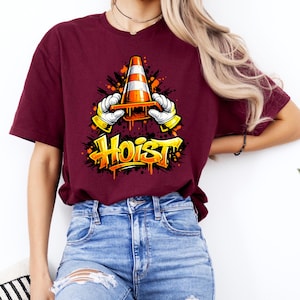 May include: Burgundy t-shirt featuring a graphic of a traffic cone held by gloved hands, with the word "HOIST" in graffiti-style lettering. The design is in orange, yellow, and black. The shirt is paired with blue jeans.