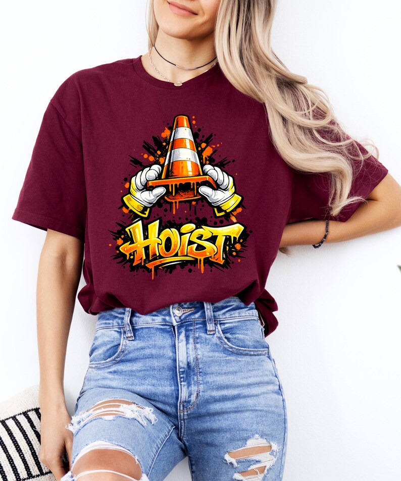 May include: Burgundy t-shirt featuring a graphic of a traffic cone held by gloved hands, with the word "HOIST" in graffiti-style lettering. The design is in orange, yellow, and black. The shirt is paired with blue jeans.