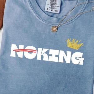 Peut inclure: T-shirt bleu clair Comfort Colors avec le mot "NOKING" en lettres blanches, barré d'un trait rouge sur "NO". Une couronne jaune est au-dessus de "KING". Un collier doré est posé sur le t-shirt.