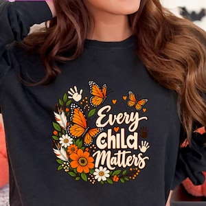 Può includere: Una felpa nera a maniche lunghe con una stampa grafica che riporta il testo Every Child Matters. Il design include farfalle monarca arancioni, fiori bianchi e arancioni, foglie verdi, piume, piccoli cuori arancioni e impronte di mani bianche.