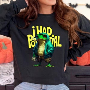 Pode incluir: Camisola preta com um sapo de desenho animado usando um capuz verde e calções. O sapo está segurando uma flor e o texto "I HAD POTENTIAL" é amarelo. Estilo casual.