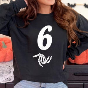 Peut inclure: Sweat-shirt noir à manches longues avec le chiffre "6" blanc et une main de squelette blanche. Vêtement décontracté, adapté à Halloween ou aux événements à thème.
