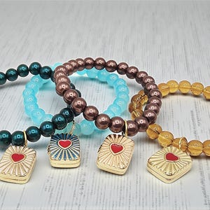 May include: Four beaded bracelets in teal, light blue, brown, and amber. Each bracelet has a gold-coloured rectangular charm with a red heart and sunburst design. The bracelets are displayed on a white surface.