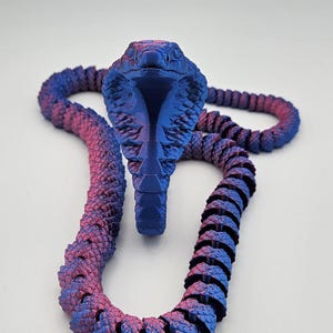 Könnte beinhalten: Ein 3D-gedrucktes Cobra-Schlangenspielzeug in Blau- und Rosatönen. Der Schlangenkopf ist aufrecht, mit einem detaillierten, strukturierten Design. Der Körper ist segmentiert, was eine flexible Bewegung ermöglicht. Der Farbverlauf sorgt für visuelles Interesse.