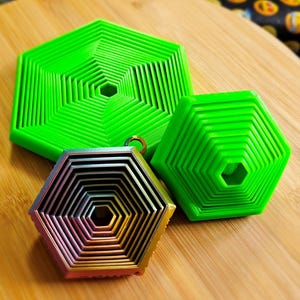 Könnte beinhalten: Drei grüne und ein schillernder mehrfarbiger sechseckiger Keksausstecher aus Metall. Die Ausstecher sind übereinander gelegt und haben ein konzentrisches Kreisdesign.