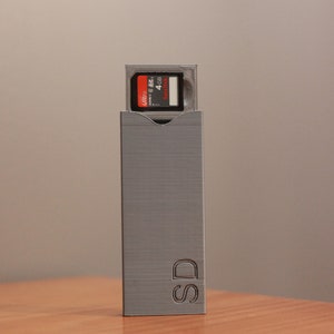 May include: A gray SD card case with a 4 GB SD card inserted. The SD card has a red and black label. The case is rectangular with the letters "SD" engraved on the side. The case is made of a textured material.