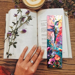 May include: A colorful bookmark featuring a hummingbird in flight with a floral design. The bookmark is made of paper and has a glossy finish.