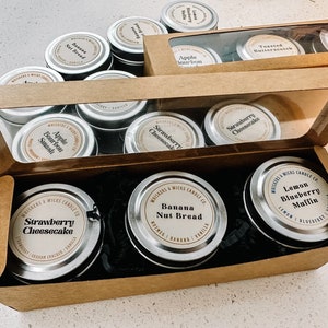 Puede incluir: Un conjunto de 10 pequeñas latas de velas redondas y plateadas con varios aromas. Los aromas incluyen pastel de queso de fresa, pan de plátano y nueces, muffin de arándanos y limón, bourbon de manzana, bourbon de manzana, pastel de queso de fresa, caramelo de mantequilla tostado, chai sucio y pera y almendra. Las velas están dispuestas en una caja marrón con una tapa transparente.