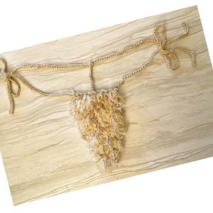 Op de afbeelding: Een beige gehaakte string met een getextureerd, gefranjerd ontwerp aan de voorkant. De string heeft gevlochten bandjes aan de zijkanten en bovenkant, wat een unieke en handgemaakte uitstraling creëert. Het item wordt gepresenteerd op een lichtgekleurd oppervlak.