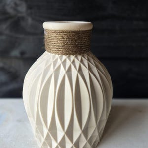 Op de afbeelding: Een crèmekleurige decoratieve vaas met een geometrisch patroon. De vaas heeft een afgeronde vorm en is versierd met een band van natuurlijk touw rond de hals. Een stijlvol woondecoratie-item.
