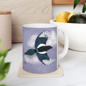 May include: White ceramic mug featuring a detailed illustration of a magpie in flight. The bird has black, white, and blue plumage against a lavender sky with clouds. The mug is on a beige coaster.