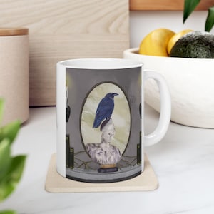 May include: White ceramic mug featuring a detailed illustration. The design includes a blue raven perched atop a classical bust within an oval frame. The background is a muted gray, with hints of books and decorative elements.