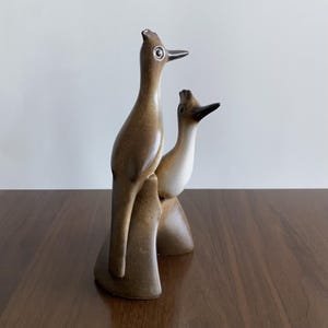 Puede incluir: Una escultura de cerámica marrón con dos pájaros estilizados posados juntos. Los pájaros tienen cuellos largos, picos y detalles en los ojos. La escultura es un artículo decorativo, adecuado para la decoración del hogar.