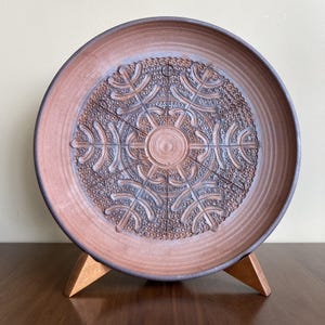 Op de afbeelding: Een bruin keramiek bord met een reliëf geometrisch ontwerp. Het bord staat op een houten standaard.