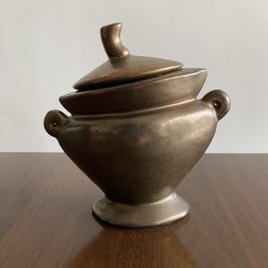 Pode incluir: Um recipiente de cerâmica de cor bronze com tampa. O recipiente tem um corpo arredondado com duas pequenas alças e uma base de pedestal. A tampa tem uma alça curva. O recipiente é provavelmente para armazenamento ou fins decorativos.