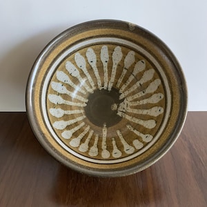 Può includere: Una ciotola in ceramica nei toni del marrone e beige. La ciotola presenta un cerchio centrale marrone scuro, circondato da un motivo di disegni bianchi a forma di cucchiaio che si irradiano verso l'esterno. Il bordo ha un bordo marrone e beige.