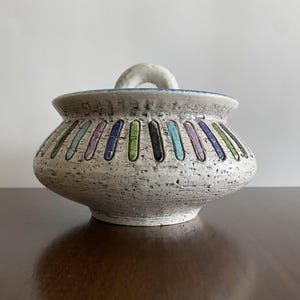 Pode incluir: Um pote de cerâmica texturizado, branco sujo, com tampa. O pote apresenta uma série de listras verticais coloridas em azul, verde, preto e roxo. A tampa tem uma alça branca arqueada e uma borda azul.