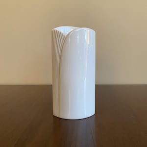 Może przedstawiać: Biały ceramiczny wazon o unikalnym, zakrzywionym wzorze. Wazon ma gładkie, błyszczące wykończenie i lekko zaokrągloną podstawę.