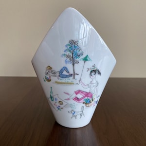 May include: White ceramic vase with a distinctive diamond shape. The vase is decorated with a colourful illustration of a picnic scene, including figures, a tree, and a dog. The design features a person reclining under a tree and a woman with a parasol.