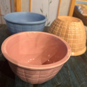 May include: Three ceramic bowls in pastel colors: light blue, pink, and tan. The pink bowl is in the foreground, with a textured, grid-like pattern. The blue bowl has a smooth surface. The tan bowl has a grid pattern.