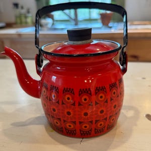 May include: A red enamel tea kettle with a black handle and lid. The kettle features a repeating pattern of orange and black floral designs. The spout is curved, and the kettle sits on a white surface.