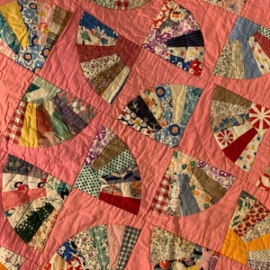 May include: Close-up of a handmade quilt featuring a fan pattern. The quilt showcases a variety of patterned fabrics in a fan-like design, set against a pink background. The fabrics include floral, geometric, and plaid patterns.