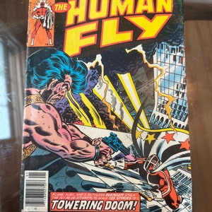 May include: Vintage comic book cover for "The Human Fly" by Marvel Comics. The cover features a muscular character with a gun, and a superhero in a red and white suit. The title is in large yellow letters, with the text "Towering Doom!"