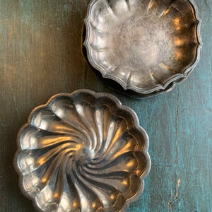 May include: A collection of antique silver-toned dishes. The dishes have a scalloped edge design. One dish has a spiral pattern in the center. The dishes are on a distressed teal-colored wooden surface.