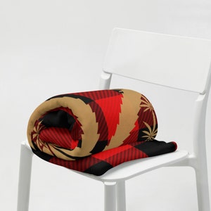 May include: A rolled up blanket with a red, black, and gold plaid pattern featuring a leaf design. The blanket is sitting on a white chair.