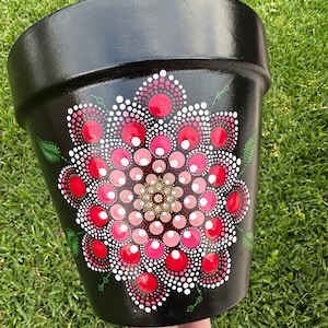 Op de afbeelding: Een zwart geschilderde bloempot met een handgeschilderd mandala-ontwerp. De mandala heeft een centraal patroon van kleine, lichtbruine stippen omgeven door concentrische cirkels van roze, rode en witte stippen. Groene bladaccenten zijn ook aanwezig.