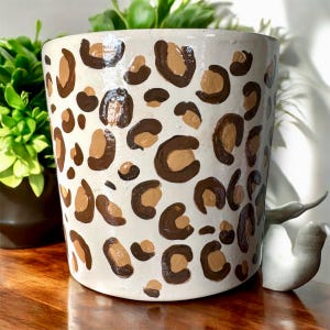 May include: A cylindrical, off-white ceramic planter with a glossy finish. The planter is decorated with a repeating pattern of brown and tan leopard spots. Green plants are visible in the background.