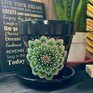 Może przedstawiać: Czarna doniczka z terakoty z namalowanym na przodzie zielono-białym wzorem mandali. Doniczka ma pasującą podstawkę.