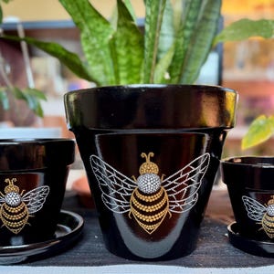 May include: Three black terracotta flower pots with gold and white bee designs. The largest pot has a large bee design with a gold body and white wings. The two smaller pots have smaller bee designs with gold bodies and white wings.