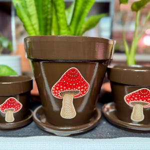 May include: Three brown ceramic flower pots of varying sizes, each adorned with a red mushroom design with white polka dots. The pots sit on matching saucers. The background features green plants.