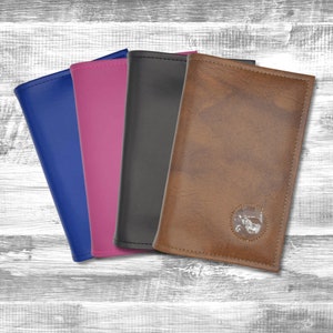 May include: Four different colored leather passport holders. The holders are all closed and stacked on top of each other. The top holder is brown with a clear window for a photo ID. The other holders are blue, pink, and black.