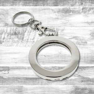 May include: A silver-colored keychain with a round, open-faced pendant. The keychain has a lobster clasp and a split ring for attaching keys. The pendant has a shiny, reflective surface and a simple, modern design.