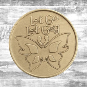 Peut inclure: Une pièce de monnaie ronde de couleur bronze avec l'inscription "Let Go, Let God" et un motif de papillon. Le fond est un motif de bois blanc et gris, créant un design simple et élégant.
