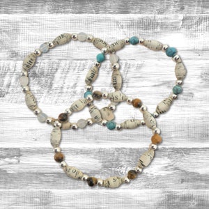 May include: Three paper bead bracelets with silver beads and turquoise and tiger's eye accents. The bracelets are intertwined and laid out on a white wood background.