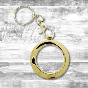 May include: Gold-toned keychain with a round, open circle charm and a swivel clasp. The keychain is on a white wooden background.